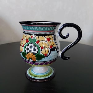 Puede incluir: Una taza de cerámica colorida con base de pedestal y asa curva. La taza presenta un vibrante diseño floral y geométrico en amarillo, verde, rojo y azul. El asa es azul oscuro y el borde es negro. La taza es probablemente hecha a mano.