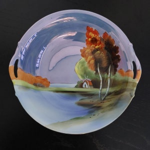 Puede incluir: Un bol de servir de porcelana vintage con una escena pintada a mano de un lago, árboles y una pequeña casa. El bol tiene dos asas y un esmalte azul claro con reflejos morados y rosados.