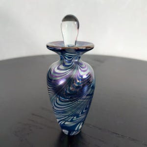 May include: A decorative glass perfume bottle with a clear stopper. The bottle features a swirling pattern of blue, silver, and purple. The bottle has a flared rim with a gold-colored edge. The stopper is clear and elongated.
