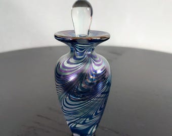 May include: A decorative glass perfume bottle with a clear stopper. The bottle features a swirling pattern of blue, silver, and purple. The bottle has a flared rim with a gold-colored edge. The stopper is clear and elongated.