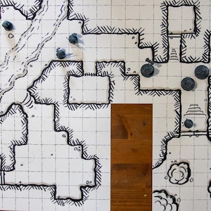May include: A hand-drawn dungeon map with a grid pattern, featuring a river, rooms, and various terrain elements. Several gray game pieces are placed on the map, suggesting a tabletop role-playing game setting. The map is on a wooden surface.