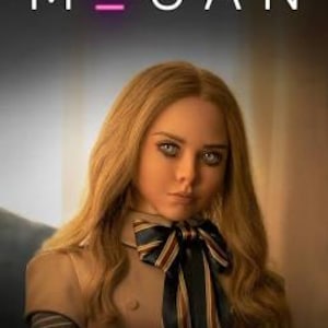 May include: A doll with long blonde hair and light eyes, wearing a tan collared dress with a dark blue, red, and white striped bow at the neck. The text M3GAN is displayed in white at the top, with the number three in bright pink.