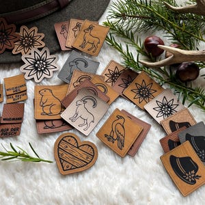 Könnte beinhalten: Eine Sammlung von Holz- und Lederaufnähern mit verschiedenen Designs, darunter Blumen, Tiere und Hüte. Einige Aufnäher haben Text. Die Aufnäher sind in Braun-, Grau- und Beigetönen gehalten und auf einer weißen, flauschigen Oberfläche angeordnet.