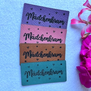 May include: Four leather labels with the text "Mädchenkram" in black lettering. The labels are in different colours: purple, pink, brown, and green.