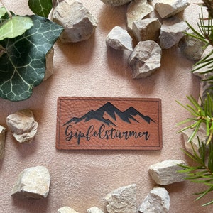 Könnte beinhalten: Ein braunes Lederpatch mit einem schwarzen Bergketten-Design und dem Text "Gipfelstürmer" in schwarzer Schrift.