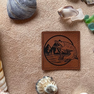 Könnte beinhalten: Ein braunes Lederstück mit einer schwarzen Silhouette von zwei Fischen, die vor einer Bergkette aus dem Wasser springen. Die Fische befinden sich in einem Kreis mit schwarzem Umriss.