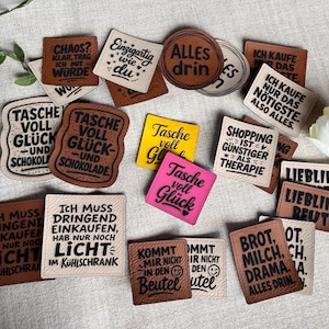 Könnte beinhalten: Eine Sammlung von Lederaufnähern in verschiedenen Formen und Farben, jeweils mit deutschem Text. Die Aufnäher zeigen Sätze wie "Chaos?" und "Shopping ist günstiger als Therapie." Einige sind quadratisch, andere rund, und einige haben dekorative Kanten.