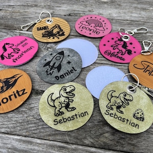 Könnte beinhalten: Eine Sammlung personalisierter Schlüsselanhänger mit verschiedenen Designs und Namen. Die Schlüsselanhänger zeigen Bilder von Dinosauriern, Raketen, Einhörnern, Fußbällen und Baggern. Die Schlüsselanhänger sind in verschiedenen Farben, darunter Rosa, Beige und Grau.
