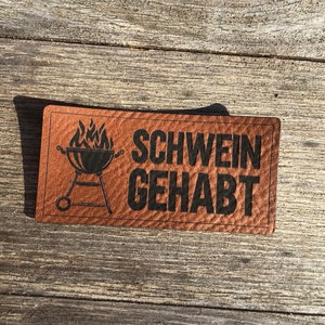 Könnte beinhalten: Ein braunes Leder-Patch mit einer schwarzen Silhouette eines Grills mit Flammen und dem Text "SCHWEIN GEHABT".