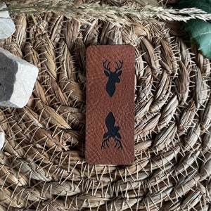 Könnte beinhalten: Ein braunes Lederstück mit einer schwarzen Silhouette eines Hirsches und einem stilisierten Baum mit Geweih. Das Stück ist auf einem geflochtenen Stroh-Hintergrund.