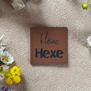 Könnte beinhalten: Ein braunes Leder-Etikett mit dem Text "kleine Hexe" in schwarzer Schrift.