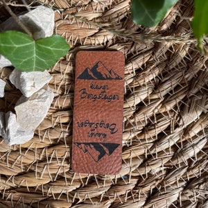 Könnte beinhalten: Ein braunes Leder-Patch mit dem Text "kleiner Bergsteiger" und einem Berg-Silhouetten-Design. Der Text ist unten auf dem Patch umgekehrt wiederholt.
