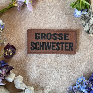 Könnte beinhalten: Ein braunes Leder-Etikett mit dem Text "GROSSE SCHWESTER" in schwarzen Buchstaben, umgeben von verschiedenen blauen und violetten Blumen.