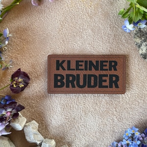 Könnte beinhalten: Ein braunes Leder-Patch mit dem Text "KLEINER BRUDER" in schwarzen Buchstaben. Das Patch ist von verschiedenen Blumen umgeben, darunter blaue Vergissmeinnicht, violette Stiefmütterchen und gelbe Löwenzahnpflanzen.