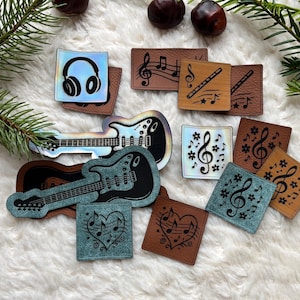 May include: Assortment of music-themed patches and guitar-shaped embellishments. Designs include headphones, musical notes, treble clefs, and a flute. The patches are in various colors, including brown, blue, and iridescent, with a white fluffy background.