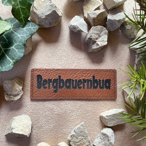 Könnte beinhalten: Ein braunes Leder-Patch mit dem Text "Bergbauernbua" in schwarzen Buchstaben. Das Patch ist umgeben von kleinen grauen Steinen und grünem Laub.