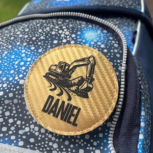 Könnte beinhalten: Nahaufnahme eines Rucksacks mit einem runden, goldfarbenen Aufnäher. Der Aufnäher zeigt ein schwarzes Bagger-Design und den Namen "DANIEL" in fetten, schwarzen Buchstaben. Der Rucksack hat ein blau-weißes Sprenkelmuster.