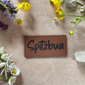 Könnte beinhalten: Ein braunes Leder-Patch mit dem Wort "Spitzbua" in schwarzer Schrift.
