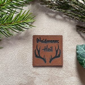 Könnte beinhalten: Ein braunes Leder-Patch mit dem Text "Waidmanns Heil" und einem Paar schwarzer Hirsche.