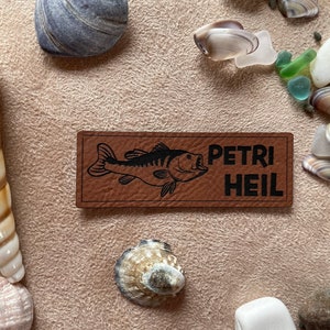Könnte beinhalten: Braunes Leder-Patch mit einer schwarzen Silhouette eines Fisches und dem Text "PETRI HEIL" in Schwarz.