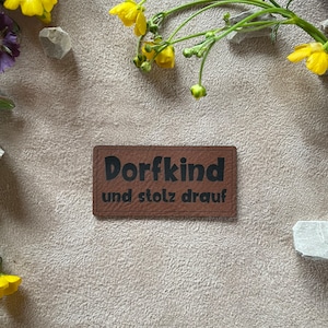 Könnte beinhalten: Ein braunes rechteckiges Patch mit den Worten "Dorfkind und stolz drauf" in schwarzer Schrift. Das Patch ist von gelben Blumen, Steinen und Grün auf einer strukturierten, beigen Oberfläche umgeben.