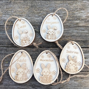 Può includere: Cinque decorazioni a forma di uovo di Pasqua in legno con i nomi Liesa, Benjamin, Nala, Maximilian e Sabrina. Ogni uovo presenta un design a forma di coniglietto tagliato al laser, fiori e uno spago di iuta. Le uova sono marroni chiare.