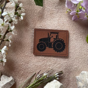 Könnte beinhalten: Eine schwarze Silhouette eines Traktors auf einem braunen Lederfleck. Der Fleck ist von weißen Blumen und grünen Blättern umgeben.