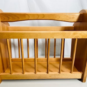 May include: A light brown wooden magazine rack featuring a rectangular base, two solid side panels with curved top edges, and a series of vertical dowels forming the front and back walls, topped with a horizontal handle bar for carrying.