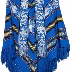May include: A bright blue poncho featuring a woven geometric pattern in white, yellow, and black. The garment includes a wide front panel with intricate snowflake-like designs, a V-neckline, and a hemline finished with long, matching blue fringe.