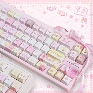 May include: A pink mechanical keyboard with white and pastel-colored keycaps. The keycaps feature various patterns, including hearts, bears, and bows. The keyboard has the text "Butterbear" and "Beckspace" on some keys. A pink ribbon is tied around the keyboard.