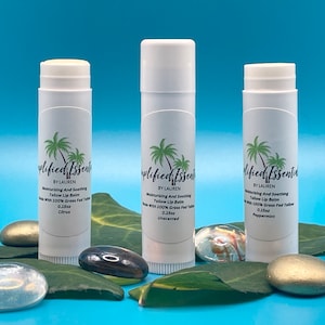 May include: Three white tubes of lip balm with green palm tree labels. The labels read "Simplified Essentials by Lauren" and list the flavors: Citrus, Unscented, and Peppermint. The tubes are sitting on green leaves and gold and gray stones against a blue background.
