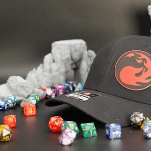 May include: A black and red baseball cap with a mesh back. The cap features a red and black logo of a stylized flame on the front. The cap is surrounded by colourful dice.