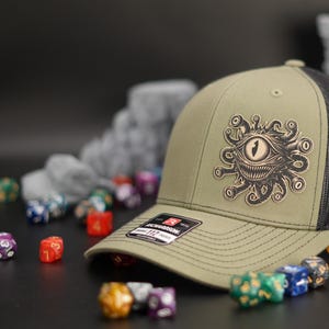 May include: A green and black trucker hat with a brown embroidered image of a beholder, a creature from the Dungeons and Dragons fantasy role-playing game. The hat is surrounded by colorful dice.