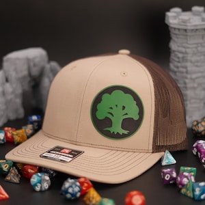 Op de afbeelding: Een bruine en beige trucker-pet met een groen boomlogo op de voorkant. Het logo is een cirkel met een groene boom erin. De pet is omringd door dobbelstenen.