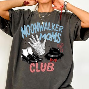 May include: Dark gray t-shirt with the text "MOONWALKER MOMS CLUB" in blue and pink. The graphic includes a white glove, black shoes, and a black hat with a red bow tie. The shirt has a vintage, worn look.
