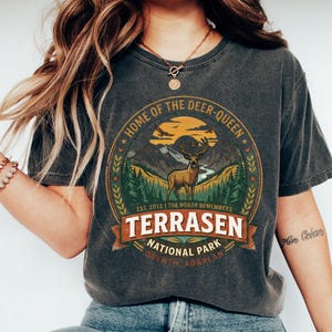 May include: Dark gray t-shirt with a circular graphic featuring a deer, mountains, and the text "HOME OF THE DEER-QUEEN" and "TERRASEN NATIONAL PARK". The design includes the text "EST. 2012 | THE NORTH REMEMBERS" and "ORYNTH ADARLAN".