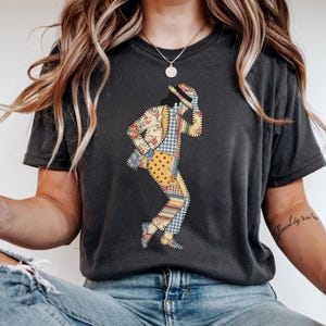 May include: Dark gray t-shirt featuring a patchwork design of a person in a dancing pose. The patchwork includes floral, polka dot, and plaid patterns. The person is wearing a hat and a suit.