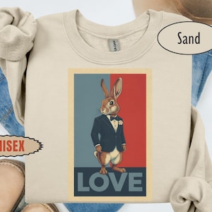 Può includere: Felpa unisex color sabbia con una grafica di un coniglio in giacca e cravatta, con la scritta "LOVE" in basso. Il coniglio è marrone e bianco, su uno sfondo rosso e blu. La felpa è sopra jeans blu e scarpe marroni.