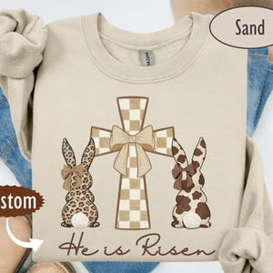 Könnte beinhalten: Ein sandfarbenes Sweatshirt mit einem Osterdesign. Das Design umfasst ein kariertes Kreuz mit einer Schleife und zwei Hasenfiguren mit gemustertem Fell. Der Text "He is Risen" ist in einer Schrift unter den Hasen und dem Kreuz geschrieben.