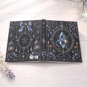 May include: An open book with a dark cover, decorated with intricate illustrations. The cover is black with silver and blue details, such as a bow, crystals, and floral patterns. Text includes "What Breaks Apart Often Reveals The Most" and "No. III PRISMATIC VEIL."