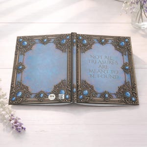 Peut inclure: Un livre relié à couverture bleue orné de cadres en filigrane couleur bronze et de fausses pierres précieuses bleues. Le côté droit affiche le texte : NOT ALL TREASURES ARE MEANT TO BE FOUND. Une petite icône de clé et un QR code sont visibles.