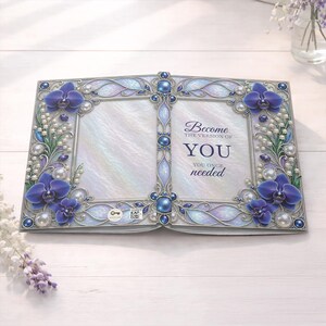 Puede incluir: Un marco de fotos decorativo abierto con un fondo nacarado. El marco está adornado con orquídeas azules, perlas y detalles plateados ornamentados. El texto "Become the version of YOU you once needed" se muestra en el lado derecho.