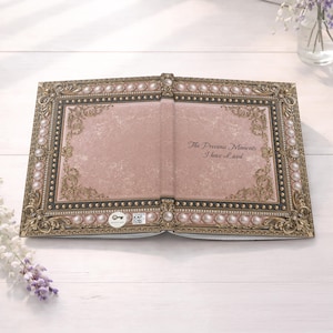 May include: An open book with a dusty rose cover featuring an ornate gold frame, a border of white pearls, and the text The Precious Moments I have Lived printed in black script. The design includes intricate gold filigree corners and studs.