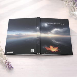 Peut inclure: Journal ouvert avec un paysage panoramique. La couverture présente un ciel dramatique au-dessus de l'eau, avec une feuille d'érable orange vif. Le texte "Custom Text Here" est visible sur la couverture droite. Le journal est ouvert sur une surface en bois blanc.
