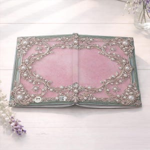 May include: Open book with a pink interior and ornate silver and pearl embellishments. The cover has a light green border and a key symbol. The design is reminiscent of vintage or antique decor.
