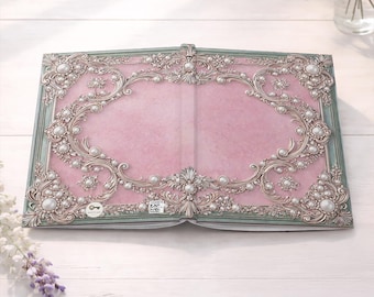May include: An open book with a pink cover featuring an ornate, raised silver frame design decorated with white pearls. The border is accented with a muted green trim. A small white circular icon with a key symbol and a QR code are visible on the cover.