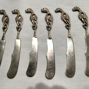 May include: Six silver butter knives with ornate dragon-shaped handles. The blades are curved and have a dull, metallic finish. The handles feature detailed dragon designs, adding a decorative touch to the set. The knives are arranged on a white surface.