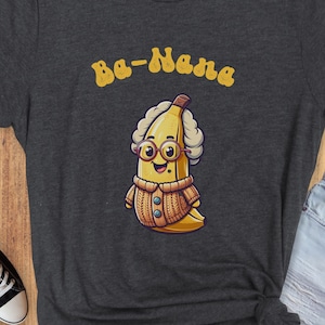 May include: A dark heather grey t-shirt with a cartoon banana wearing glasses and a sweater. The text "Ba-Nana" is printed in yellow above the banana.