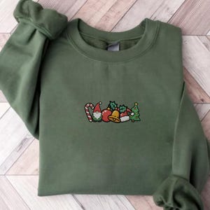 Puede incluir: Sudadera verde militar con diseño navideño bordado. El diseño incluye un bastón de caramelo, un gnomo, adornos, acebo, una campana y un árbol de Navidad. El texto "Green Military" está en la parte inferior.