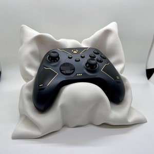 May include: A black and gold Xbox controller with a white pillow underneath. The controller has a textured design and gold accents.